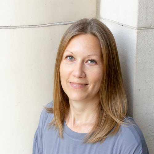 A woman with straight, shoulder-length blonde hair and a light gray shirt smiles softly while standing against a light-colored wall, reflecting the calm ambiance often found at Bowen therapy in Utah County.